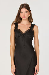 A woman is wearing a sleek black slip dress with lace detailing on the shoulder straps and neckline. The dress has a smooth, shiny fabric and a slim fit that extends below the waist. She accessorizes with gold hoop earrings, a gold bracelet, and rings on both hands.